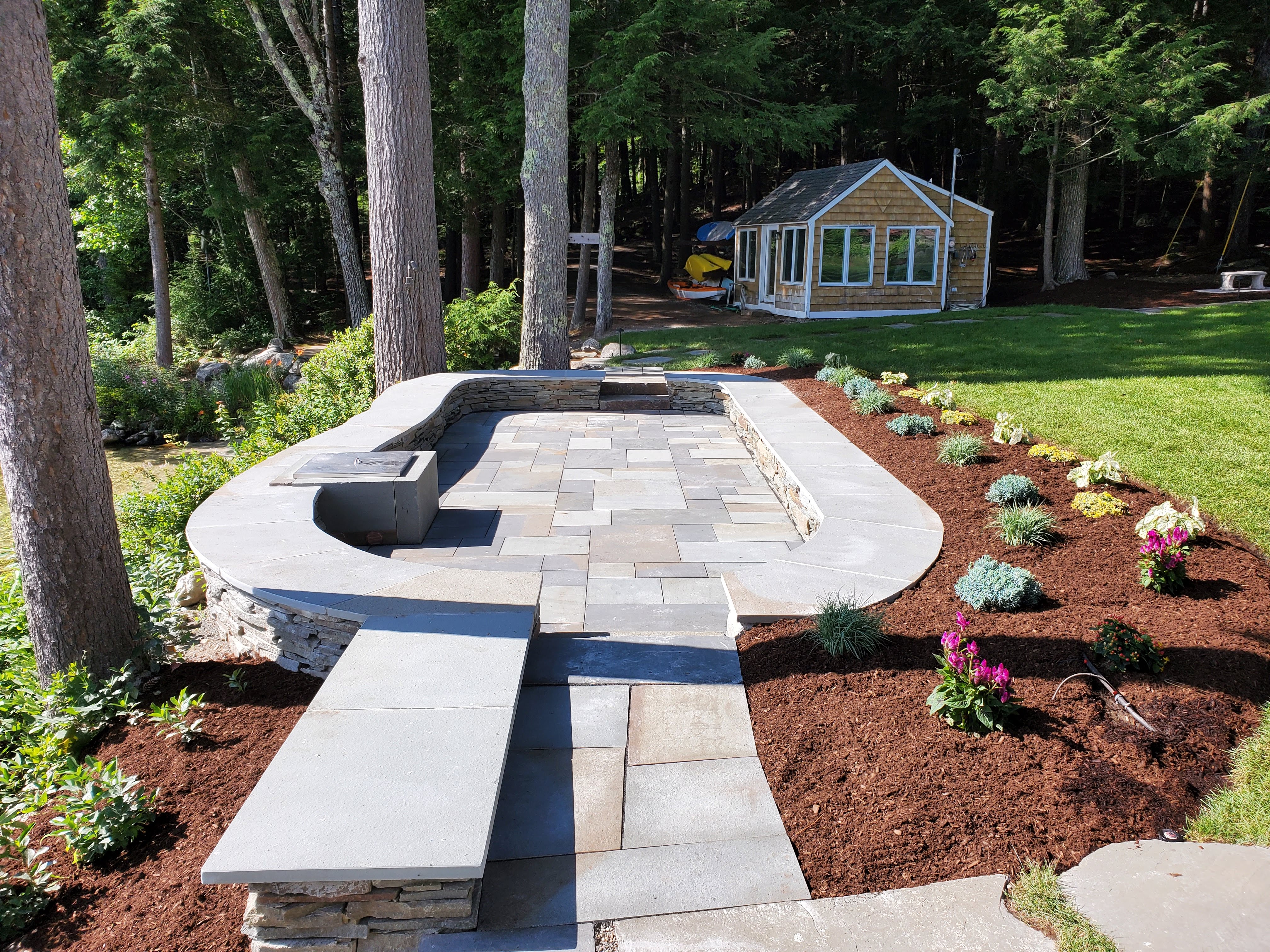 Finished bluestone patio with fire pit nook and stone seat wall in a wooded setting