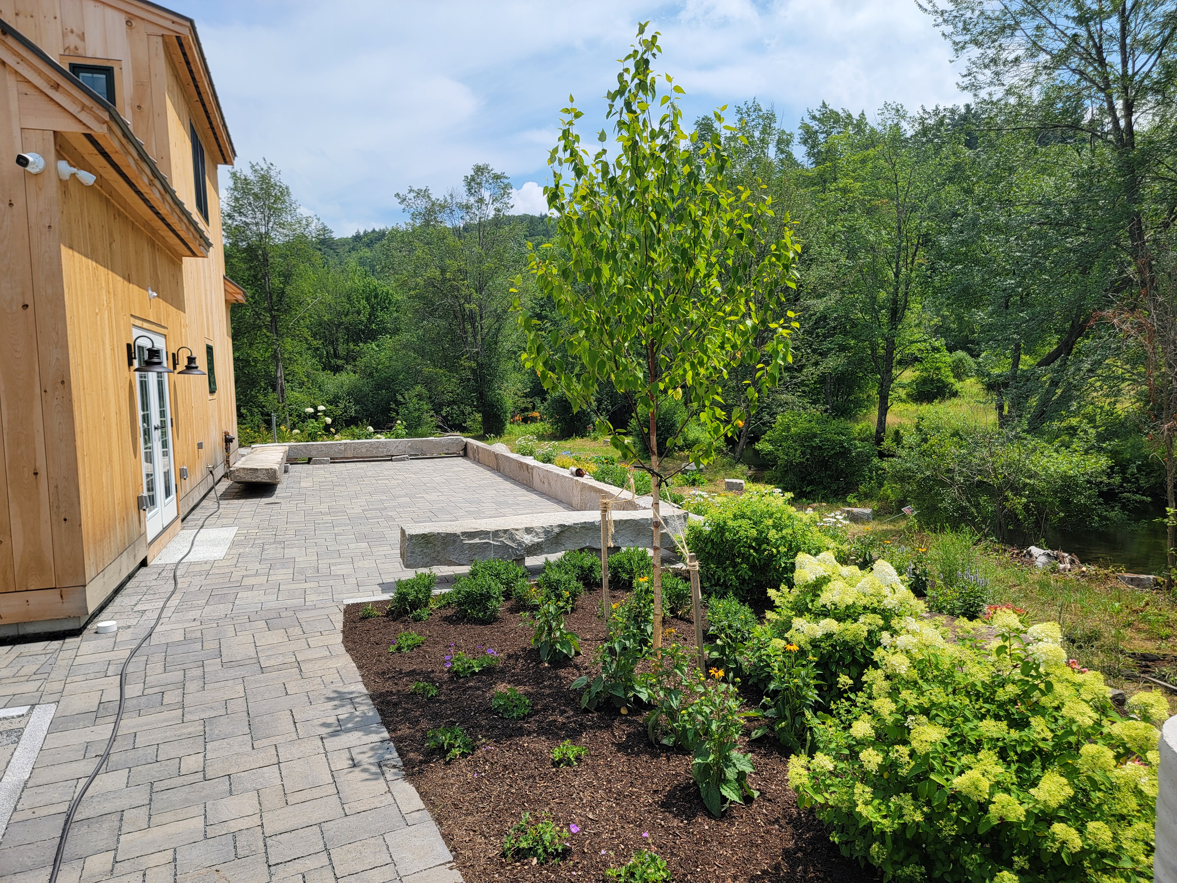Brick paver patio next to a wood-clad outbuilding with new perennial bed