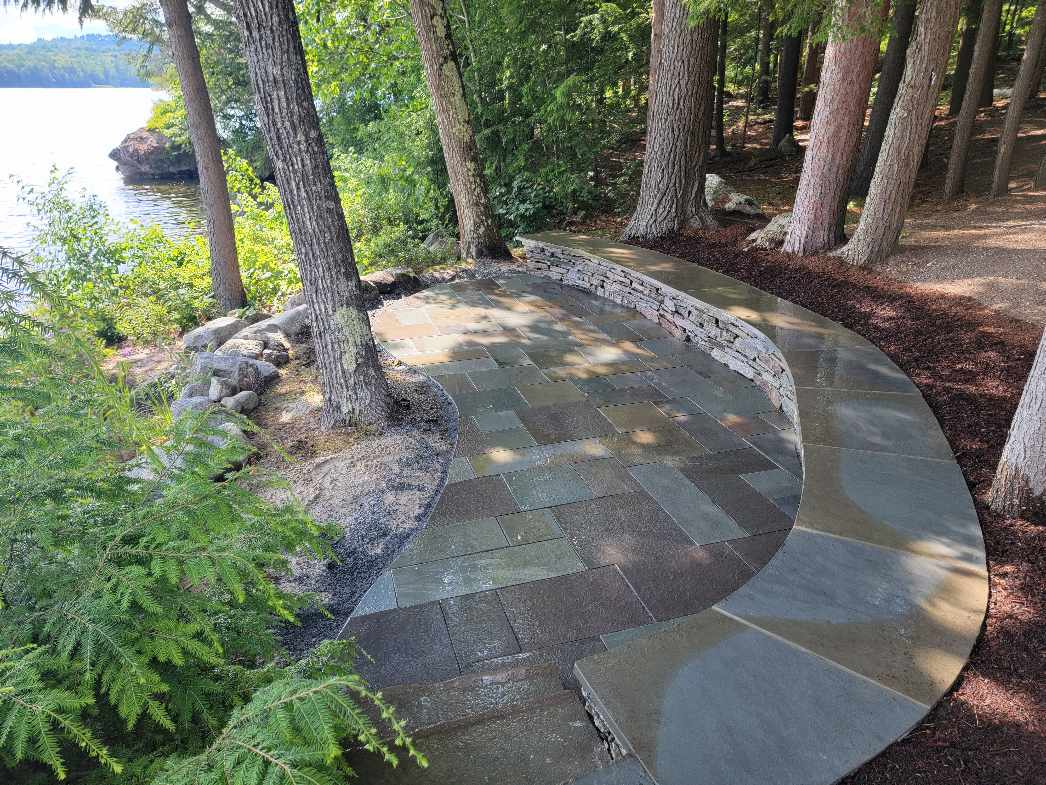 Curved bluestone patio with low stacked-stone retaining wall, lake glimpsed through pines