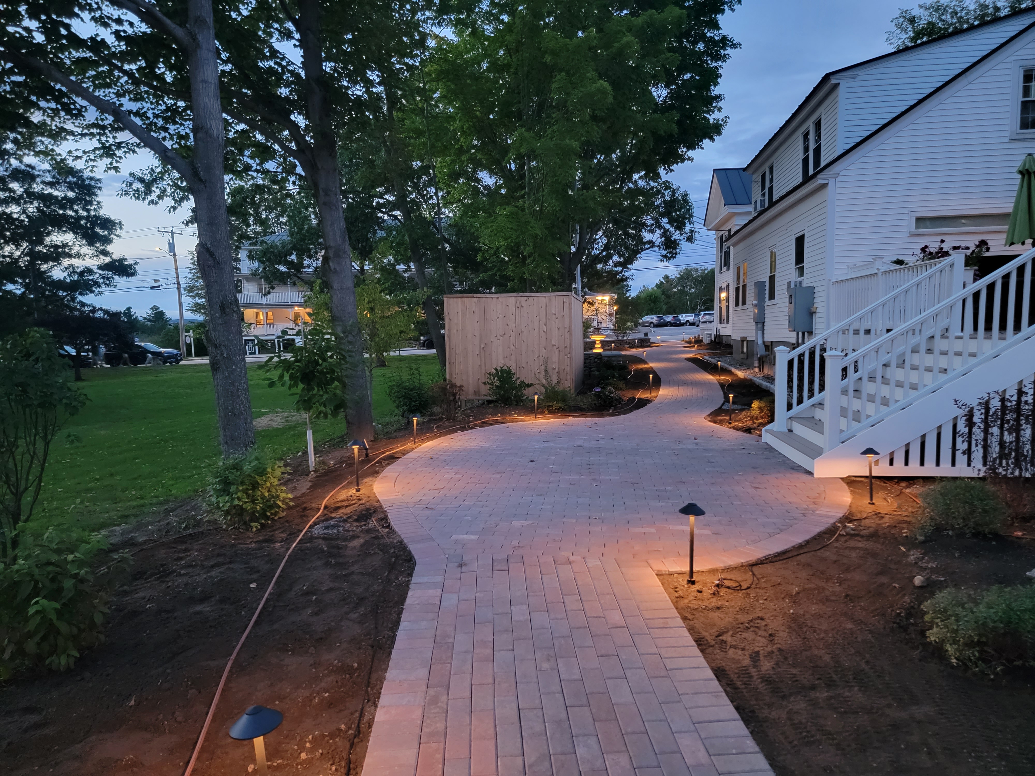 Brick walkway curving past a clapboard home, illuminated at dusk