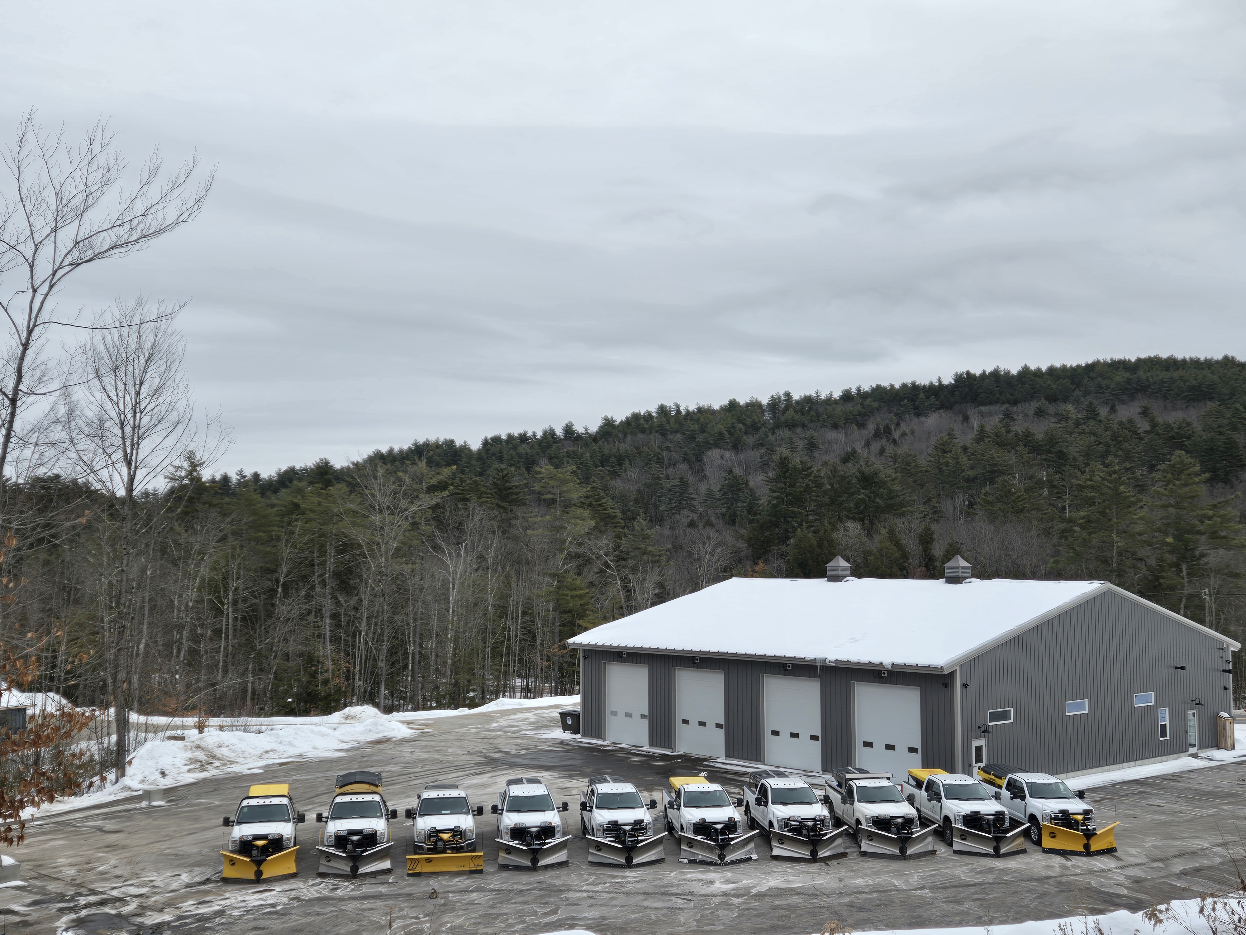 Fleet of plow trucks lined up at the company shop with snowy hills behind