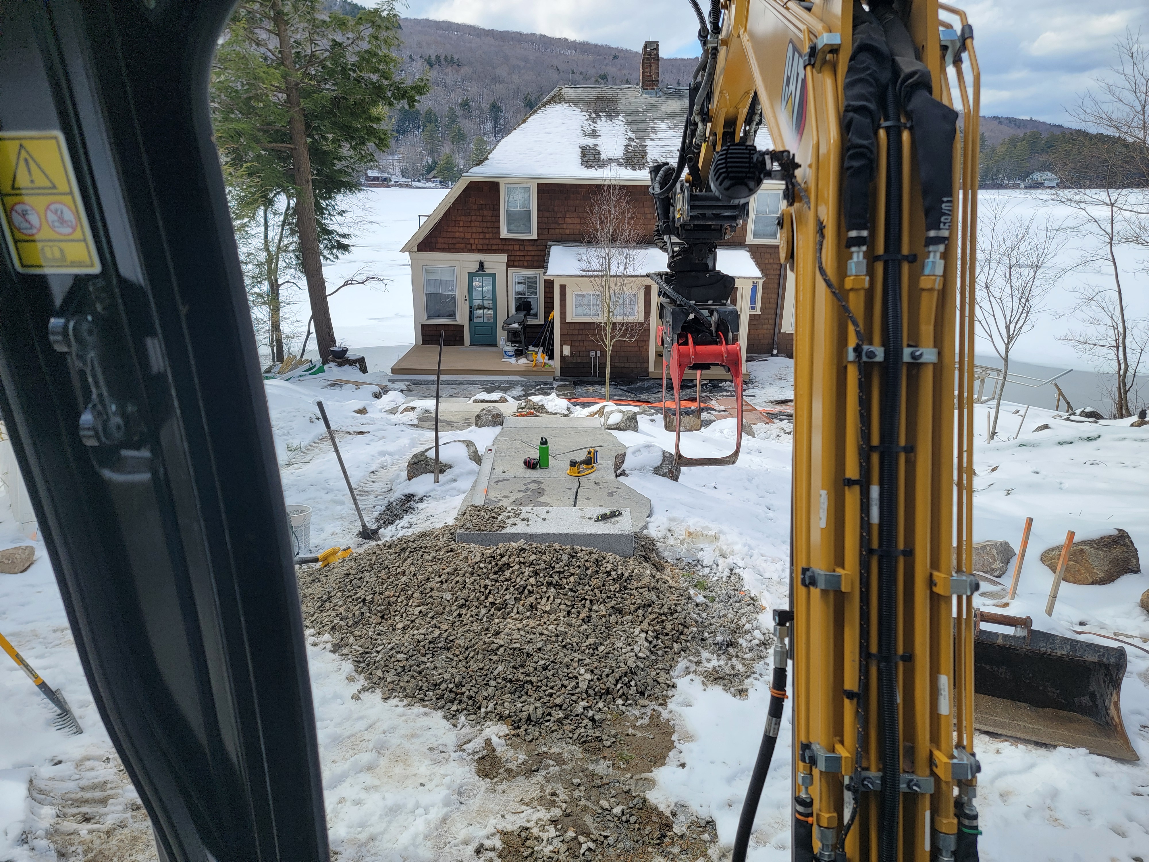 Winter shot from inside an excavator cab toward a snow-covered lakefront cottage